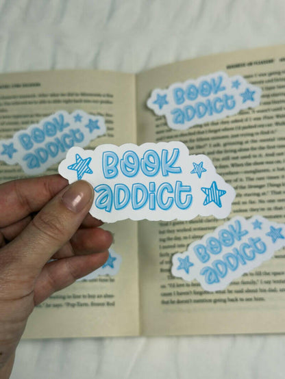 Sticker met de tekst 'Book Addict' in speelse blauwe letters met sterretjes, perfect voor boekenliefhebbers.